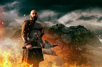 Angry viking with axe dressed in traditional nordic clothes standing in fire, battle in rocky mountains. Scandinavian ancient warrior