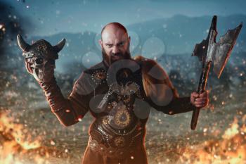 Angry viking dressed in traditional nordic clothes holds axe and human skull, battle in fire. Scandinavian ancient warrior