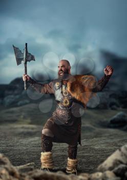 Viking with axe dressed in traditional nordic clothes standing at the rocky mountains and raised his hands up, barbarian. Scandinavian ancient warrior