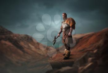 Handsome viking with axe dressed in traditional nordic clothes standing on the top of rocky mountain. Scandinavian ancient warrior