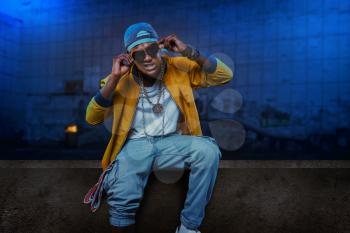 Black rapper in cap and sunglasses poses in underpass, singer in grunge subway. Rap performer on scene with lights, underground music, urban style