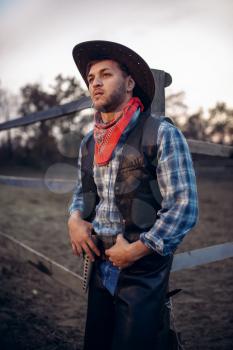Young cowboy in leather jacket and hat poses against horse corral, western. Vintage male person, retro american fashion in wild west style