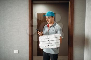 Pizza delivery boy in t-shirt and cap calls to customer, delivering service. Courier from pizzeria holds cardboard packages and phone indoors
