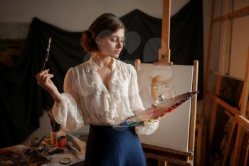 Female artist with color palette and brush standing against easel in studio. Creative paint, painter drawing portrait, workshop interior