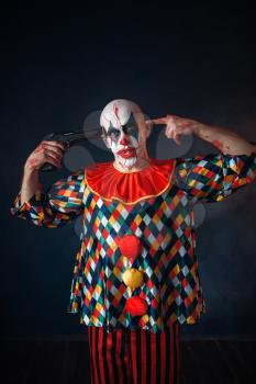 Mad bloody clown with a gun in his head, circus horror. Man with makeup in carnival costume, crazy maniac