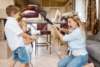 Housewife with kids playing vacuum cleaner and hair dryer at the ironing board. Woman with child doing housework at home together. Female person with daughter having fun in their house