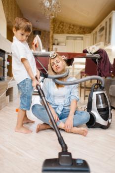 A little boy wrapped the housewife with a hose from the vacuum cleaner. Woman doing housework at home. Female person with her playful son fooling around in the house
