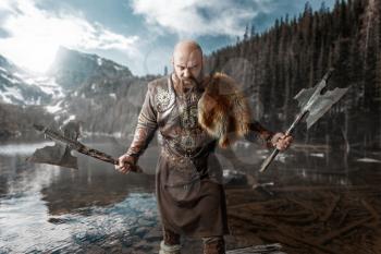 Viking with axes in hands dressed in traditional nordic clothes standing at the lake. Ancient warrior at the river, rocky mountains on background