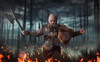 Angry viking with axe dressed in traditional nordic clothes fighting in fire, battle in forest. Scandinavian ancient warrior
