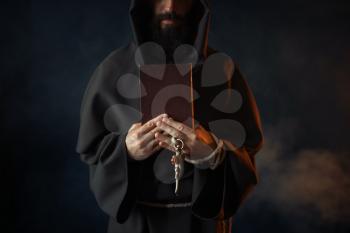 Medieval monk holds book and wooden cross in hands, secret ritual. Mysterious friar in dark cape. Mystery and spirituality