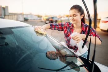 Cute woman cleans front glass of the car with sponge and spray, carwash. Lady on self-service automobile washing. Outdoor vehicle cleaning