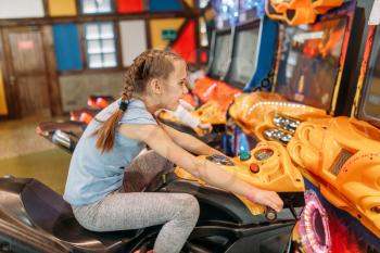 Two little girls plays game machine, children entertainment center. Excited childs having fun on playground indoors. Kids playing on motorcycles, amusement centre