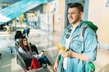 Male tourist with backpack holds burger, female travelers waiting for departure in airport on background. Passengers with baggage in air terminal, happy journey, summer travel
