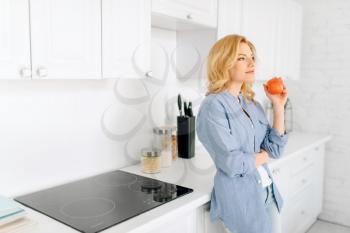 Portrait of woman with apple poses on the kitchen with snow-white interior. Female person at home in the morning, healthy nutrition and lifestyle