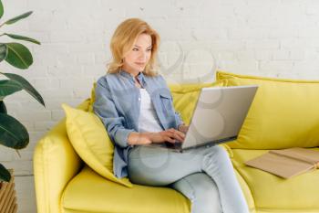 Young lady reading a book on cozy yellow couch, living room in white tones on background. Attractive female person with magazine sitting on sofa at home
