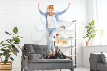 Funny woman jumping on black couch, living room in white tones on background. Cheerful female person having fun on sofa at home
