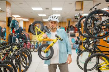 Man in cycling helmet holds children's bicycle, shopping in sports shop. Summer season extreme lifestyle, active leisure store, customer buying cycle and equipment for family riding