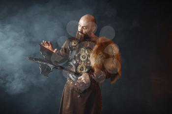 Bearded viking dressed in traditional nordic clothes touches the axe blade, barbarian image. Ancient warrior in smoke on dark background