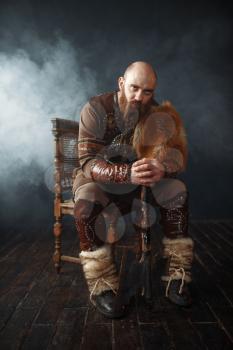 Bearded viking with axe dressed in traditional nordic clothes sitting on chair, barbarian image. Ancient warrior in smoke on dark background