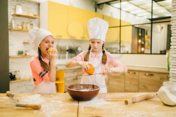 Two little girls cooks in caps rubs orange to the bowl, cookies preparation on the kitchen. Kids cooking pastry, children chefs preparing cake