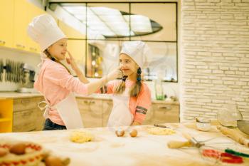Two smiling little girls cooks in caps and aprons having fun, cookies preparation on the kitchen. Kids cooking pastry, children chefs preparing cake