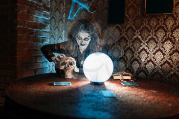 Scary witch with human skull reads a magic spell over a crystal ball, young people hands up on spiritual seance. Female foreteller calls the spirits