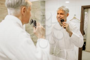 Cheerful adult man in bathrobe shaves at the mirror in the bathroom. Grey-haired mature male person with electric shaver