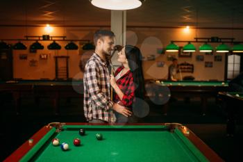 Couple hugs at the table with colorful balls in billiard room. Man and woman plays american pool game in sport bar