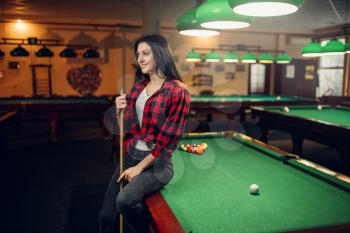 Female billiard player with cue poses at the table with colorful balls. Woman plays american pool game in sport bar