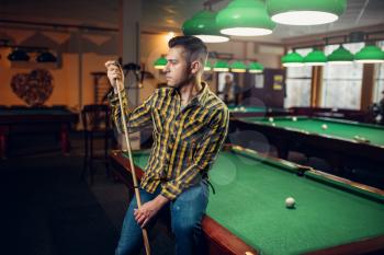 Male billiard player with cue poses at the green table. Man plays american pool game in sport bar