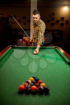 Male billiard player with cue aiming at the table with colorful balls. Man plays american game in the sport bar
