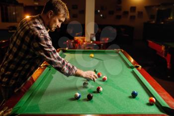 Male billiard player places balls on green table, poolroom interior on background. Man plays american pool game in sport bar