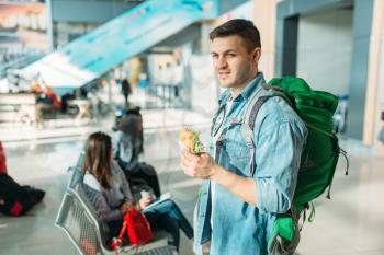 Male tourist with backpack holds burger, female travelers waiting for departure in airport on background. Passengers with baggage in air terminal, happy journey, summer travel
