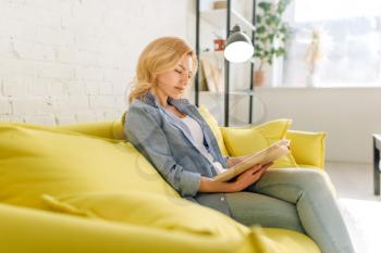Young woman reading a book on cozy yellow couch, living room in white tones on background. Attractive female person with magazine sitting on sofa