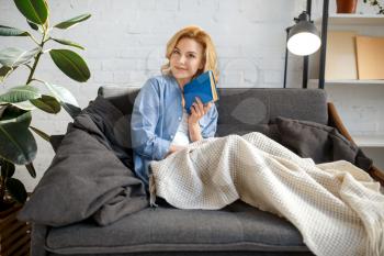 Young woman under a blanket reading a book on cozy yellow couch, living room in white tones on background. Attractive female person with magazine sitting on sofa