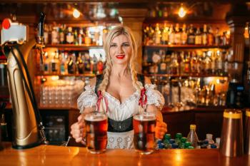 Sexy waitress holds two mugs of fresh beer at the counter in pub. Octoberfest barmaid with attractive shapes in traditional style dress