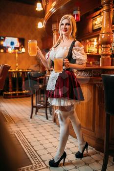 Sexy waitress in retro uniform holds two mugs of fresh beer in pub. Octoberfest barmaid with attractive shapes in traditional style dress