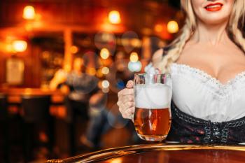 Large breasted waitress  holds mug of fresh beer in pub. Octoberfest barmaid with attractive shapes in traditional style dress. Seductive woman serving table in bar