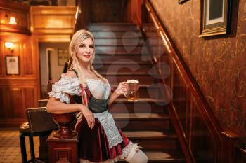 Sexy waitress with mug of fresh beer on the stairway in vintage pub. Octoberfest barmaid with attractive shapes in traditional style dress. Seductive woman holds glass of foamy beverage