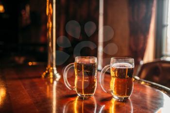 Two beer mugs on wooden bar counter, macro, nobody. Octoberfest symbol or concept. Glasses with golden beverage and foam on the table in pub, closeup view, blur background