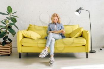 Young woman reading a book on cozy yellow couch, living room in white tones on background. Attractive female person with magazine sitting on sofa at home