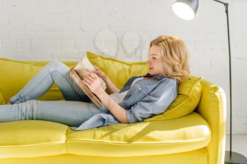 Young woman lying on cozy yellow couch and reading a book, living room in white tones on background. Attractive female person with magazine sitting on sofa