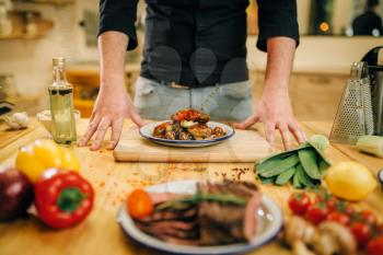 Plate wit roasted meat slices on wooden table, Male chef and fresh cooked dish for gourmets, kitchen on background. Man preparing beef with vegetables on countertop