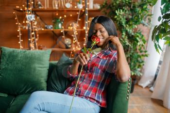 Young black woman with red rose sitting on sofa, romantic meeting at home. Happy african american female person on the couch in living room