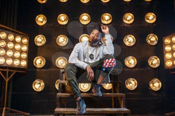 Black rapper in cap poses, perfomance on stage with spotlights on background. Rap performer on scene with lights, underground music, urban style
