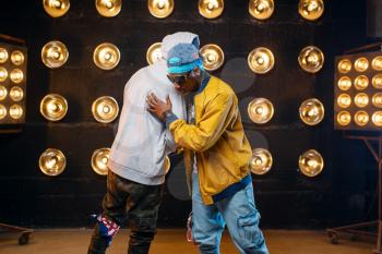 Two black rappers in caps hugging on stage, perfomance in club with spotlights on background. Rap performers on scene with lights, underground music concert, urban style
