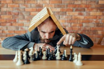 Male chess player with board on his head looking on figures, thinking process. Chessplayer playing on intellectual tournament indoors. Chessboard on wooden table, strategy game