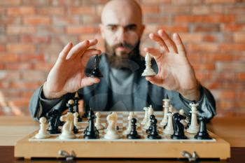 Male chess player holds white and black figures, front view. Chessplayer at board, intellectual tournament indoors. Chessboard on wooden table, strategy game