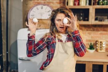 Playful housewife in an apron covered her eyes with eggs, kitchen interior on background. Female cook prepares fresh homemade cake. Domestic pie preparation