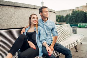 Young love couple sitting on the bench in summer city park. Smiling teenagers poses together, cityscape on background